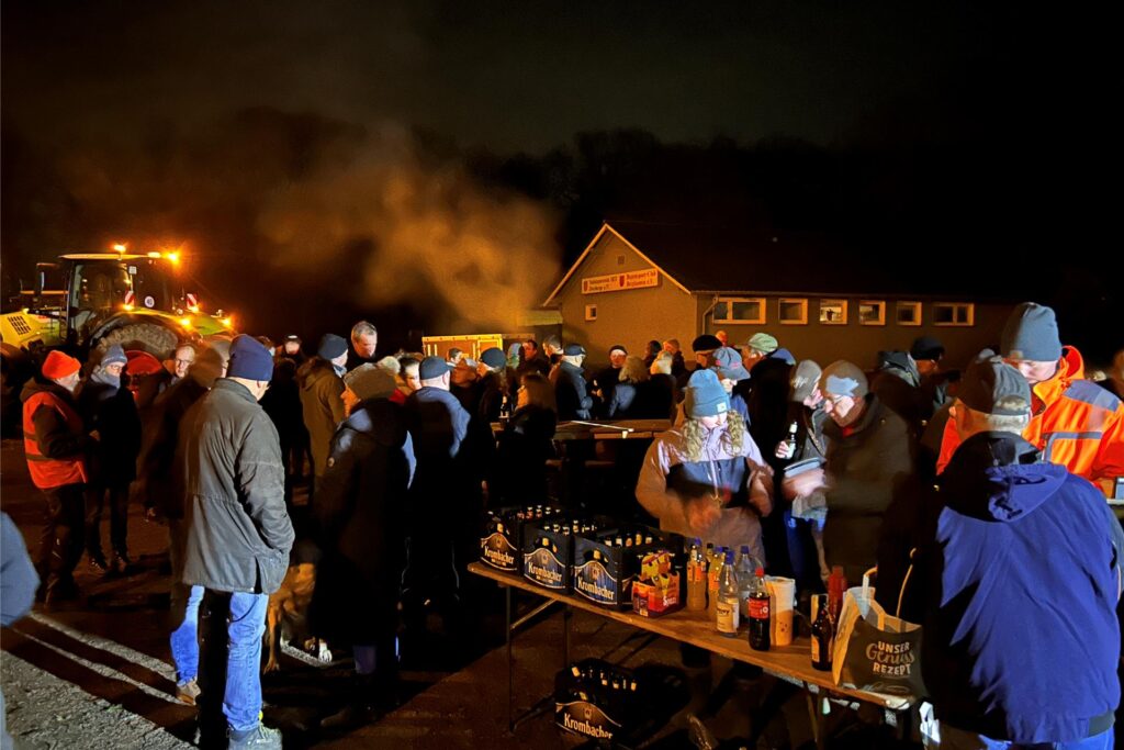 Menschen stehen in der Dunkelheit draußen an langen Tischen, auf denen Speisen und Getränke stehen.
