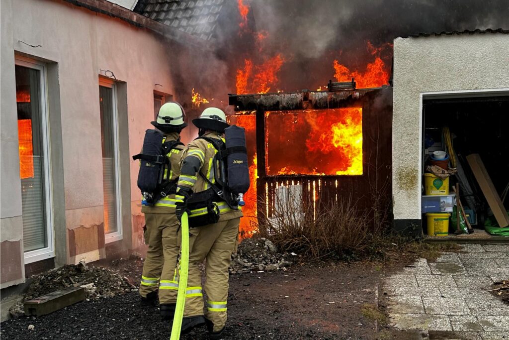 Zwei Feuerwehrleute löschen mit einem Wasserschlauch einen brennenden Anbau in Menden. Zwei Feuerwehrleute löschen mit einem Wasserschlauch einen brennenden Anbau in Menden.