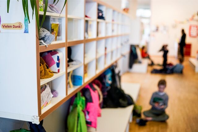 In einer Kindertagesstätte liegen verschiedene Habseligkeiten in einem Regal. Im Hintergrund spielt ein Kind auf dem Boden.