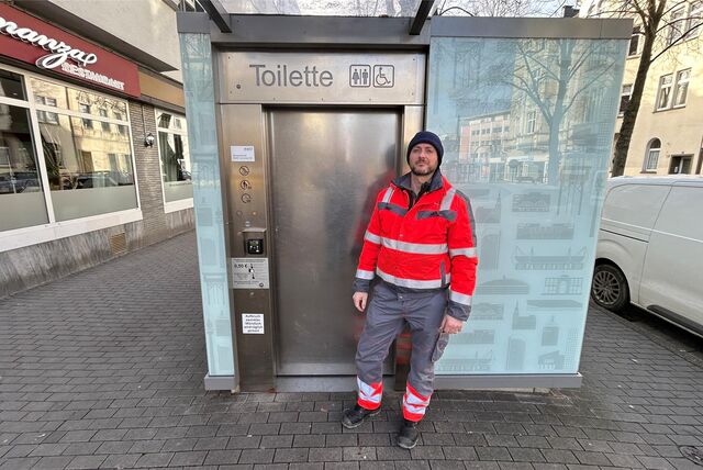 Dimitrios Sardis steht in einer orangen Arbeitsjacke vor der City-Toilette an der Kaiserstraße 99.