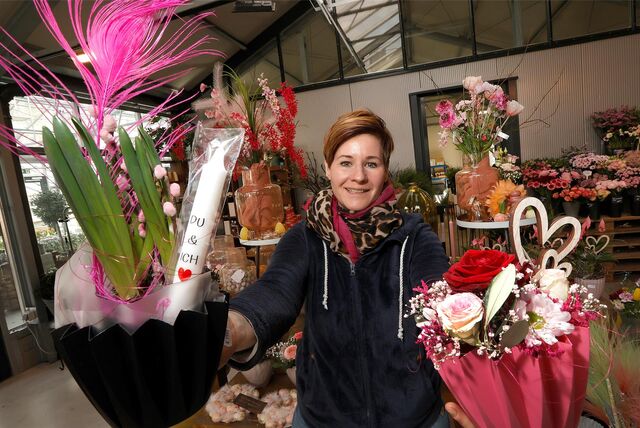 Eine Floristin, die zwei Blumengestecke für den Valentinstag in Richtung Kamera hält. Es wird viel mit Herzen, Pink und Rosa gearbeitet. Dazu sind Rosen zu sehen. Im Hintergrund stehen weitere Blumen.