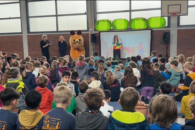 Viele Kinder in einer Turnhalle schauen zu einer Frau, die vor einer Leinwand steht. Neben ihr steht ein Mausmaskottchen.