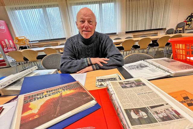 Ein älterer Mann mit Halbglatze und grauen Haaren sitzt mit verschränkten Armen an einem Tisch. Vor ihm viele Zeitungsartikel und Fotos seiner Laufbahn als Feuerwehrmann.