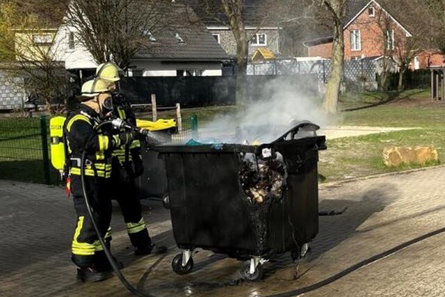 Zwei Feuerwehrleute mit Atemschutzgeräten löschen den Abfallcontainer.