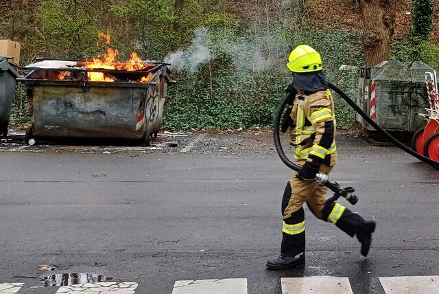 Ein Feuerwehrmann zieht den Lösch-Schlauch vom Feuerwehrwagen in Richtung Feuer. Ein großer Papiercontainer brennt.