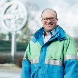 Ein Mann in blau-grün-grauer Windjacke steht am Bayerwerk in Bergkamen. Im Hintergrund sieht man das „Bayer“-Logo.