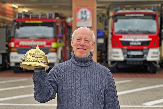 Ein älterer Mann mit Halbglatze und grauen Haaren hält einen goldenen Helm hoch. Er trägt einen blau-gauen Strickpullover. Im Hintergrund stehen zwei große Feuerwehrautos in der Garage.