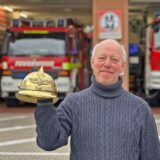 Ein älterer Mann mit Halbglatze und grauen Haaren hält einen goldenen Helm hoch. Er trägt einen blau-gauen Strickpullover. Im Hintergrund stehen zwei große Feuerwehrautos in der Garage.