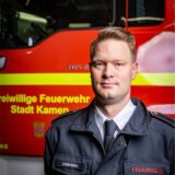 Im Hintergrund steht ein Feuerwehrwagen, auf dem „Freiwillige Feuerwehr Stadt Kamen“ steht. Im Vordergrund das Porträt eines Mannes in Dienstuniform. Er hat blonde Harre mit Seitenscheitel, blaue Augen und schaut in die Kamera.