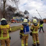 Am Rande des Großbrands in Herten-Süd besprechen sich drei Einsatzkräfte der Feuerwehr Recklinghausen. Sie blicken zu dem brennenden Gebäude, aus dem dunkler Rauch aufsteigt.