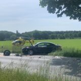 Ein Fahrzeug steht demoliert am Straßenrand. Reifen und Trümmerteile liegen verstreut auf der Fahrbahn. Ein Rettungshubschrauber steht dahinter auf einer Wiese.
