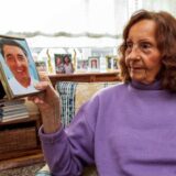 Karin Assauer sitzt in einem Sessel und hält ein Porträtfoto ihres Zwillingsbruders Rudi in Händen. Sie blickt auf die Aufnahme, die in einem kleinen Rahmen steckt.