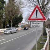 An der Straße Büddenberg in Unna weist ein Warnschild auf Straßenschäden hin. Zwei Autos fahren über die Straße. Auf beiden Seiten stehen Absperrungen.