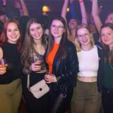 Fünf junge Frauen auf einer Party. Im hintergrund werfen Menschen enthusiastisch die Hände in die Luft.