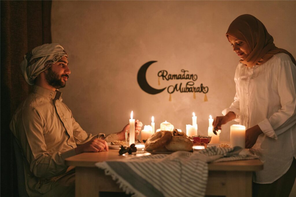 Ein Muslim sitzt an einem mit Kerzen erleuchteten Tisch, während eine Muslima Speisen für das Fastenbrechen (Iftar) während des Ramadan drauf stellt. Ein Muslim sitzt an einem mit Kerzen erleuchteten Tisch, während eine Muslima Speisen für das Fastenbrechen (Iftar) während des Ramadan drauf stellt.