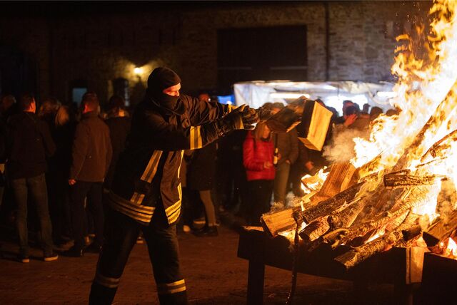Ein Feuerwehrmann mit Brandschutzhaube auf dem Kopf wirft zwei Holzscheite auf ein großes loderndes Osterfeuer. Im Hintergrund stehen viele Menschen um sich das Feuer anzuschauen.