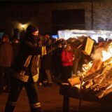 Ein Feuerwehrmann mit Brandschutzhaube auf dem Kopf wirft zwei Holzscheite auf ein großes loderndes Osterfeuer. Im Hintergrund stehen viele Menschen um sich das Feuer anzuschauen.