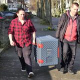 Eine Frau und ein Mann tragen eine Box mit Essen über einen Bürgersteig. Sie sind Aktive des Vereins Helping Hands in Unna, der Bedürftige und Obdachlose unterstützt.