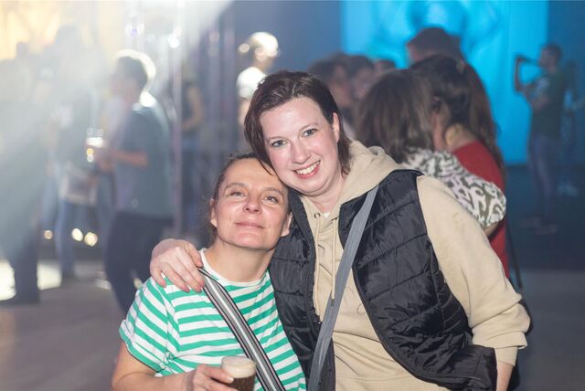 Zwei Frauen halten gegenseitig die Arme um die Schultern gelegt und lachen in die Kamera, eine hält ein Getränk in der Hand.