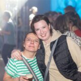 Zwei Frauen halten gegenseitig die Arme um die Schultern gelegt und lachen in die Kamera, eine hält ein Getränk in der Hand.