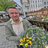 Ein junger Mann steht vor einem Blumengesteck aus Stiefmütterchen und Löwenmäulchen. Im Hintergrund ein Schild: „Kamen blüht auf 27. – 28. April“.