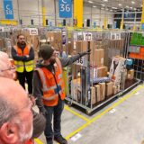 Im Verteilzentrum Unna erklärt ein Amazon-Mitarbeiter die Abläufe. Er zeigt auf Wagen voller Pakete, die in mehreren Reihen stehen.