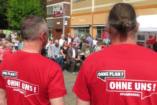 Zwei Männer mit roten T-Shirts und der Aufschrift „Ohne Plan? Ohne uns!“ auf dem Rücken stehen bei einer Maikundgebung auf dem Platz der Kulturen in Unna