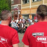 Zwei Männer mit roten T-Shirts und der Aufschrift „Ohne Plan? Ohne uns!“ auf dem Rücken stehen bei einer Maikundgebung auf dem Platz der Kulturen in Unna