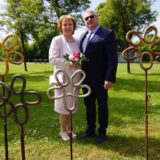 Das Brautpaar steht auf einer Wiese, während metallene Blumen des Kettenschmiedemuseums im Vordergrund zu sehen sind.