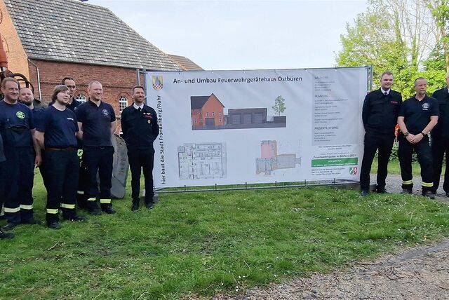Feuerwehrleute stehen an einem Banner, das den geplanten Feuerwehr-Neubau in Ostbüren abbildet.