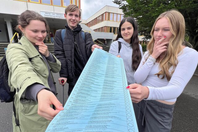 Drei Schülerinnen und ein Schüler blicken nachdenklich auf einen Musterwahlzettel für die Europawahl.