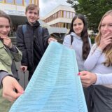 Drei Schülerinnen und ein Schüler blicken nachdenklich auf einen Musterwahlzettel für die Europawahl.