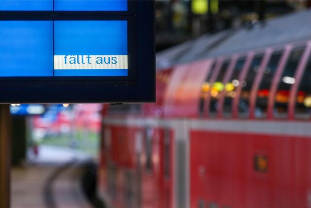 Im Hintergrund ist verschwommen ein roter Zug an einem Bahnsteig zu sehen. Im Vordergrund steht auf einer blauen Anzeigetafel „fällt aus“.