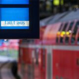 Im Hintergrund ist verschwommen ein roter Zug an einem Bahnsteig zu sehen. Im Vordergrund steht auf einer blauen Anzeigetafel „fällt aus“.