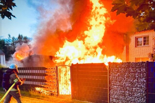 Ein Feuerwehrmann in voller Montur steht vor einer brennenden Gartenlaube.