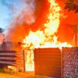 Ein Feuerwehrmann in voller Montur steht vor einer brennenden Gartenlaube.