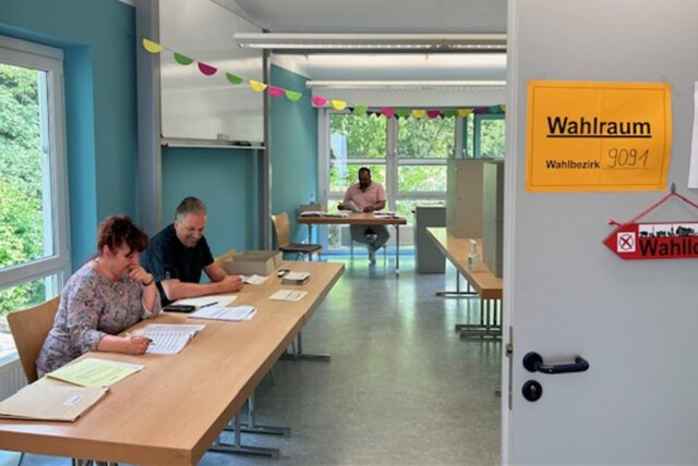 Im Klassenraum einer Schule ist ein Wahlraum eingerichtet. Das Bild zeigt den Wahlvorstand mit zahlreichen Dokumenten vor sich an einem langen Tisch.