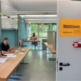 Im Klassenraum einer Schule ist ein Wahlraum eingerichtet. Das Bild zeigt den Wahlvorstand mit zahlreichen Dokumenten vor sich an einem langen Tisch.