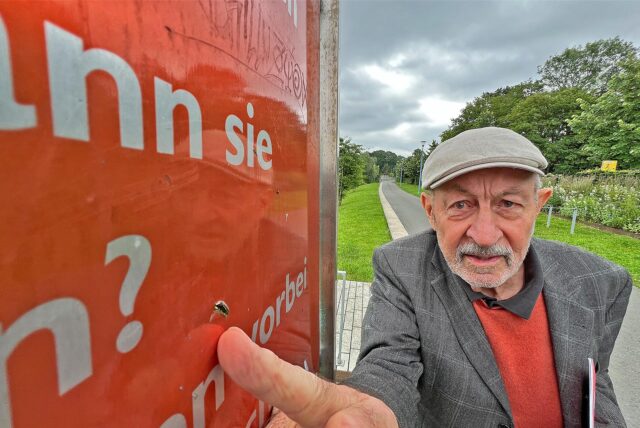 Ortsheimatpfleger Klaus Holzer zeigt mit dem Finger auf eine Tafel des Literaturpfads im Sesekepark in Kamen.