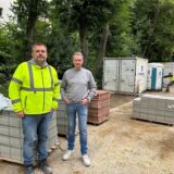 Zwei Männer stehen auf einer Baustelle. Im Hintergrund Container, Backsteine und ein Dixi-Klo.