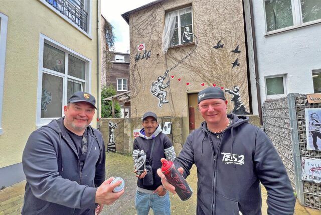 Drei Männer stehen auf einer Hauswand. Zwei halten eine Spraydose in der Hand. Im Hintergrund ist das mit Graffitibild an der Hauswand zu sehen.