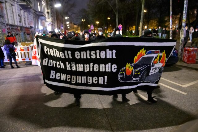 Menschen halten bei einer Demonstration ein Banner hoch, auf dem steht: „Freiheit entsteht durch kämpfende Bewegungen“. Daneben ist das Bild eines brennenden Polizeifahrzeugs zu sehen.
