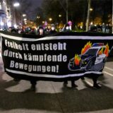 Menschen halten bei einer Demonstration ein Banner hoch, auf dem steht: „Freiheit entsteht durch kämpfende Bewegungen“. Daneben ist das Bild eines brennenden Polizeifahrzeugs zu sehen.