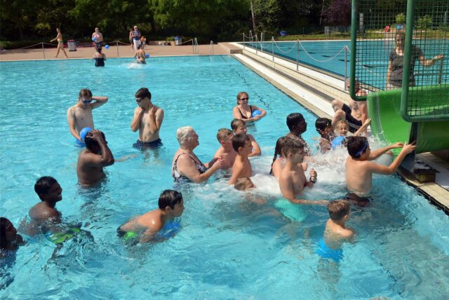 Menschen schwimmen im Freibadwasser und sind vor einer grünen Rutsche versammelt.