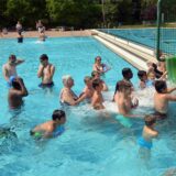 Menschen schwimmen im Freibadwasser und sind vor einer grünen Rutsche versammelt.