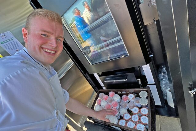 Zu sehen ist ein junger Mann, der einen Eisautomaten befüllt und in die Kamera lacht.