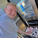 Zu sehen ist ein junger Mann, der einen Eisautomaten befüllt und in die Kamera lacht.
