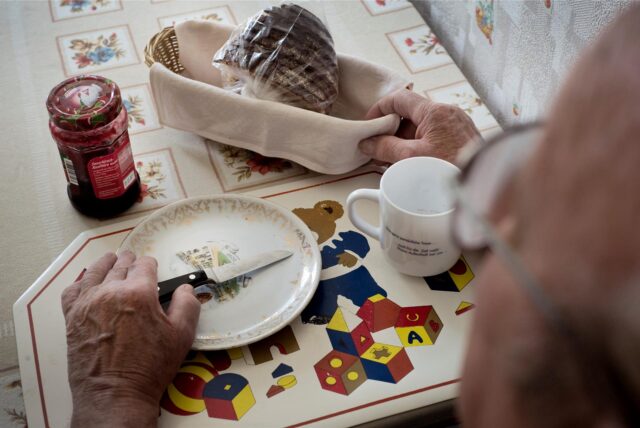 Ein älterer Mann sitzt an einem Tisch. Darauf stehen ein leerer Teller, ein Glas Marmelade, ein Brotkorb und eine Tasse
