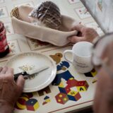Ein älterer Mann sitzt an einem Tisch. Darauf stehen ein leerer Teller, ein Glas Marmelade, ein Brotkorb und eine Tasse