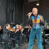 Ein Sänger in bunter Weste steht auf einer Bühne und singt ins Mikrofon.